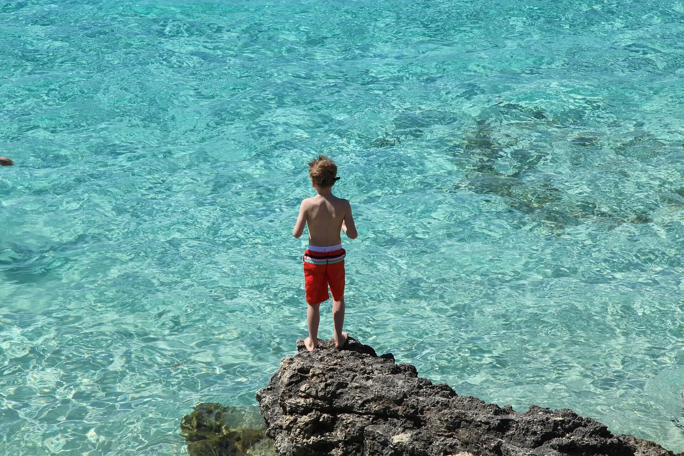 Niño de pie sobre una roca frente al agua turquesa y transparente de la Laguna Azul de Comino