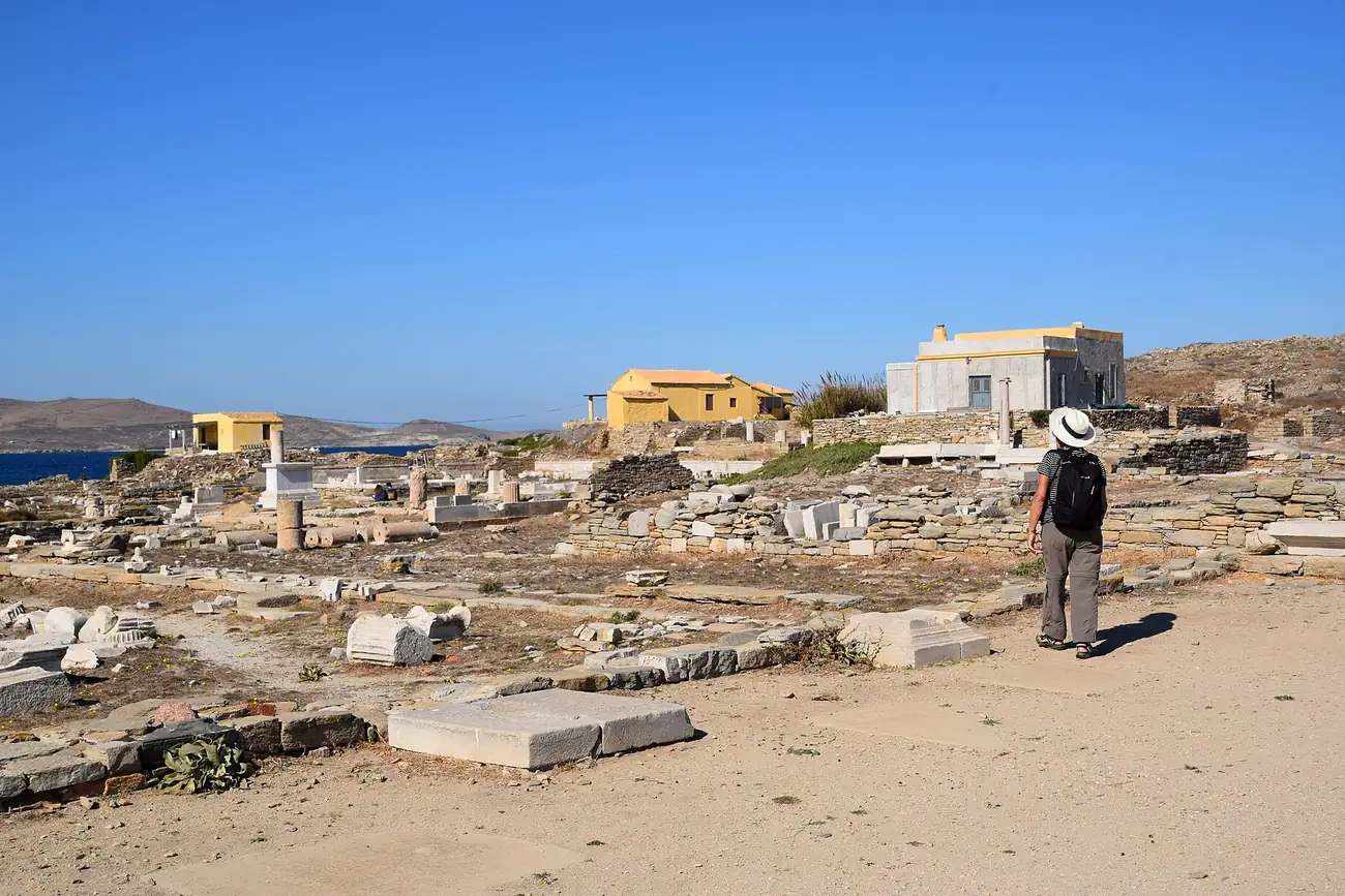 Visitante con sombrero recorriendo las ruinas del yacimiento de Delos a pleno sol, con el mar al fondo